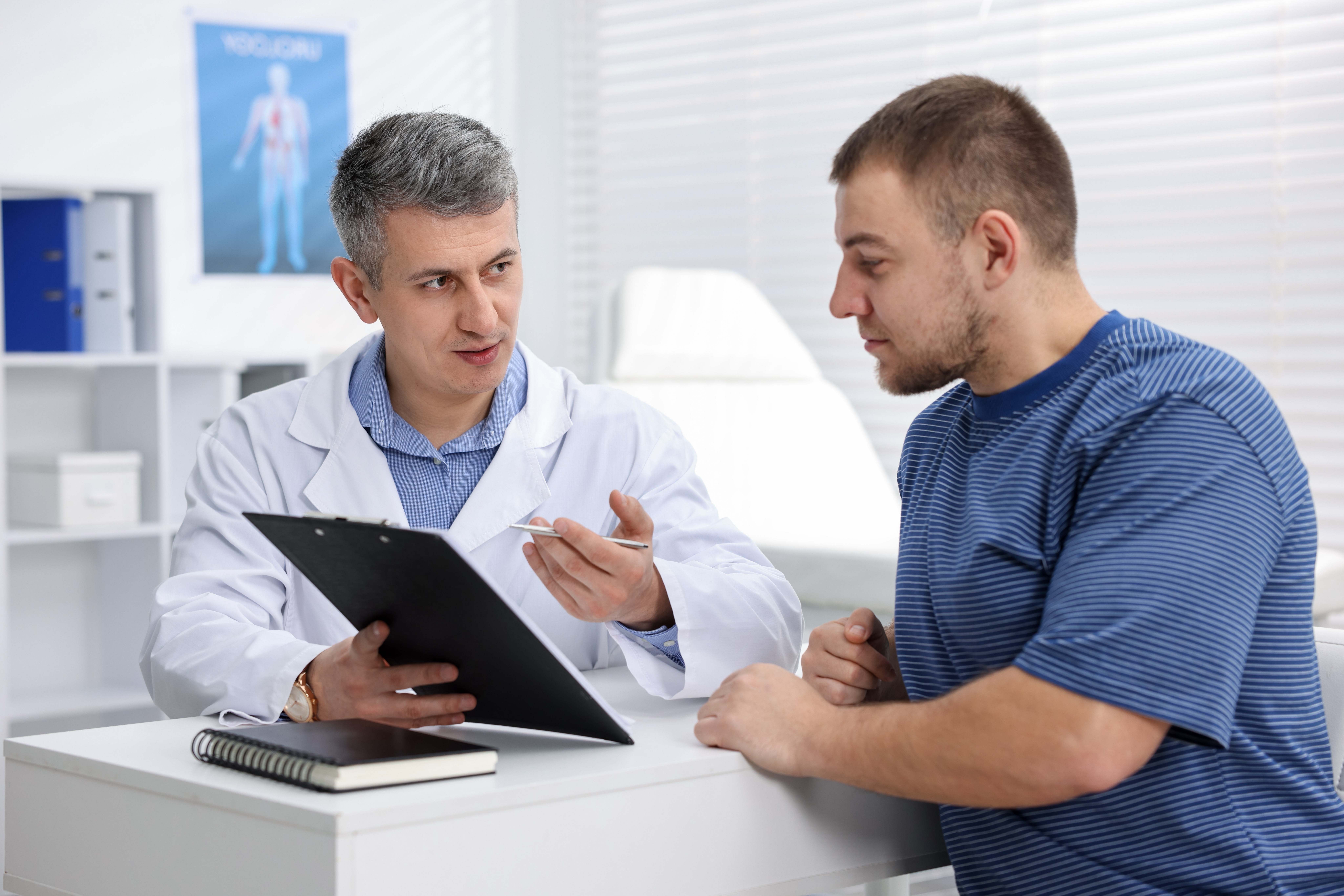 urologist,showing,clipboard,to,patient,at,white,desk,in,clinic