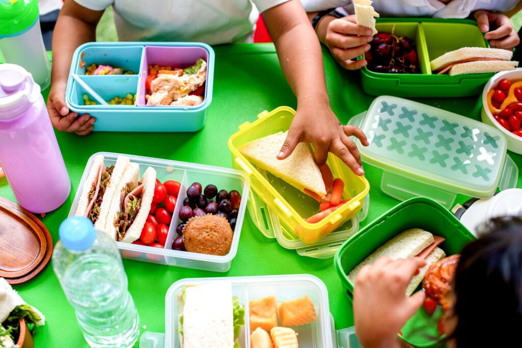 children's,hands,reaching,for,colorful,lunchboxes,filled,with,sandwiches,,fruits,